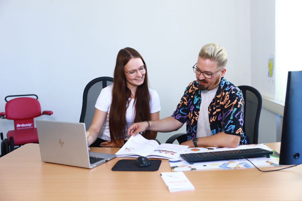 Praktikantin sitzt mit einem Mitarbeiter am Schreibtisch. Sie schauen sich einen Katalog von Rollstuhl Br&auml;unig an.