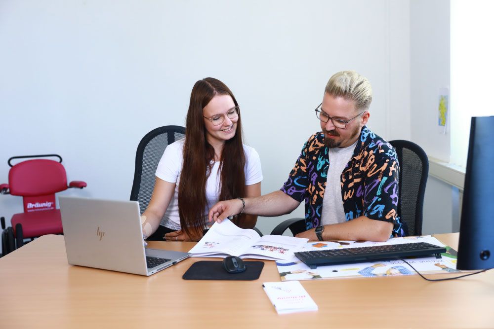 Praktikantin sitzt mit einem Mitarbeiter am Schreibtisch. Sie schauen sich einen Katalog von Rollstuhl Bräunig an.