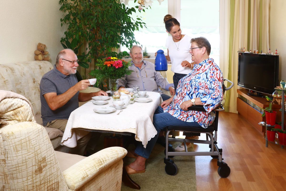 Frau im Rollstuhl sitzt an einer Kaffeetafel in ihrer engen Wohnung mit drei Gästen